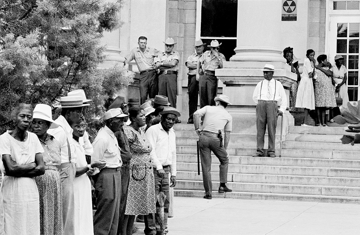 People are lined up outside of the Leflore County Courthouse in Greenwood, Mississippi, on July 16, 1964, waiting to register to vote. Several police officers stand on the steps of the courthouse.