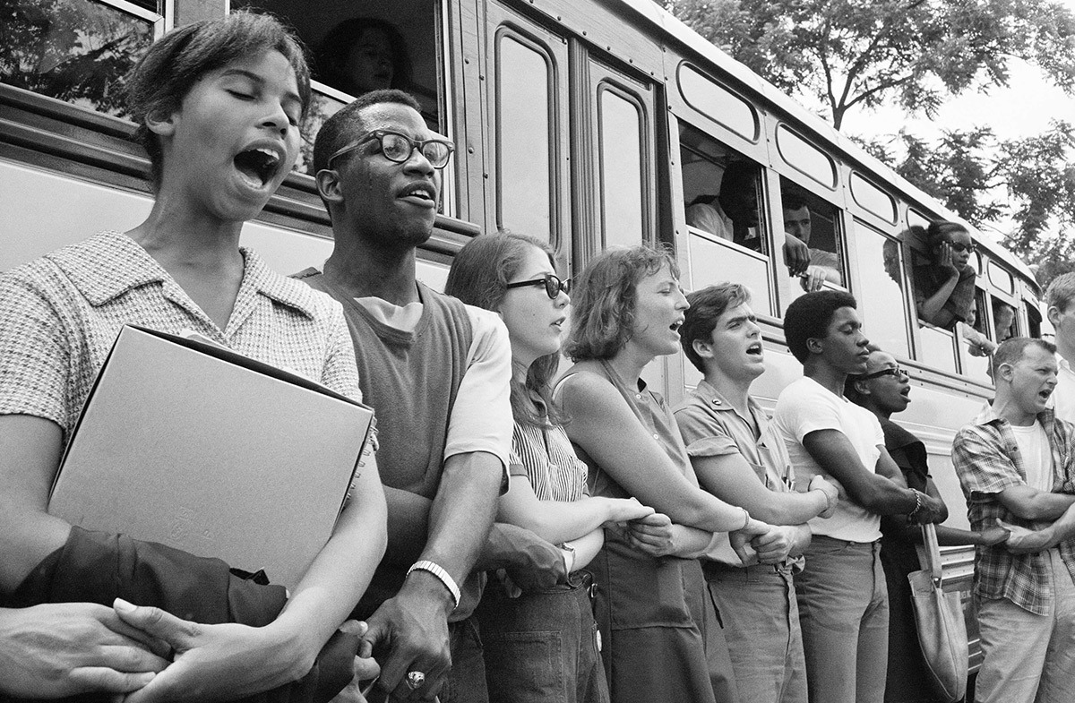 Freedom Summer 1964 volunteers stand hand-in-hand beside their bus, singing together before leaving for Mississippi.