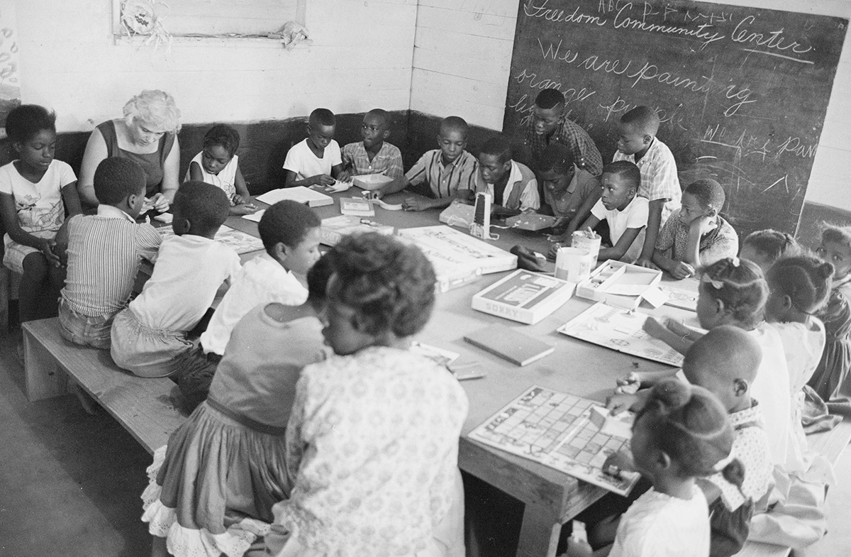 About a dozen young Black children sit around a large table with Black and white volunteer teachers. Behind them is a chalkboard wall.