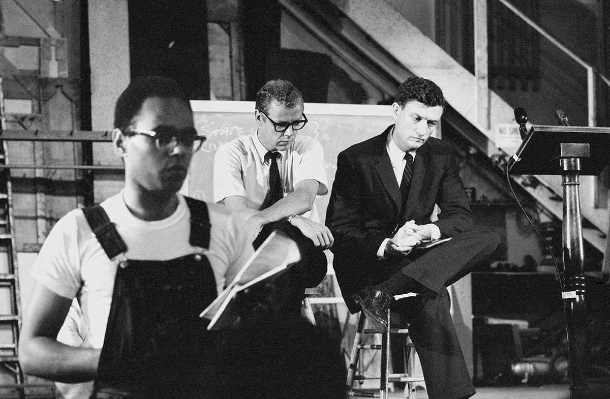 Bob Moses wears overalls and glasses while reading aloud from a paper in an auditorium. Two men seated on stools include John Doar who wears a suit and looks serious.