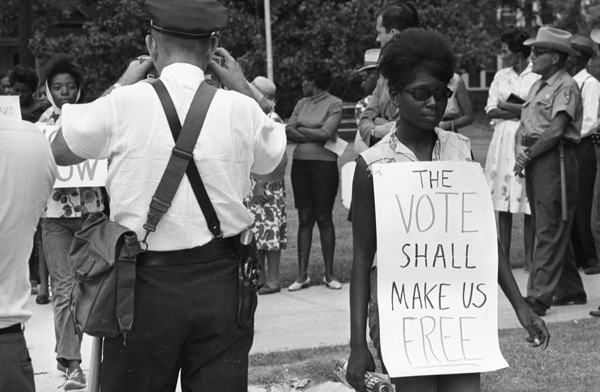 Protesters stand outside of the Leflore County Courthouse in Greenwood, Mississippi, on July 16, 1964. A woman holds a picket sign that reads “The Vote Shall Make Us Free,” while a police officer takes photos of the protesters.