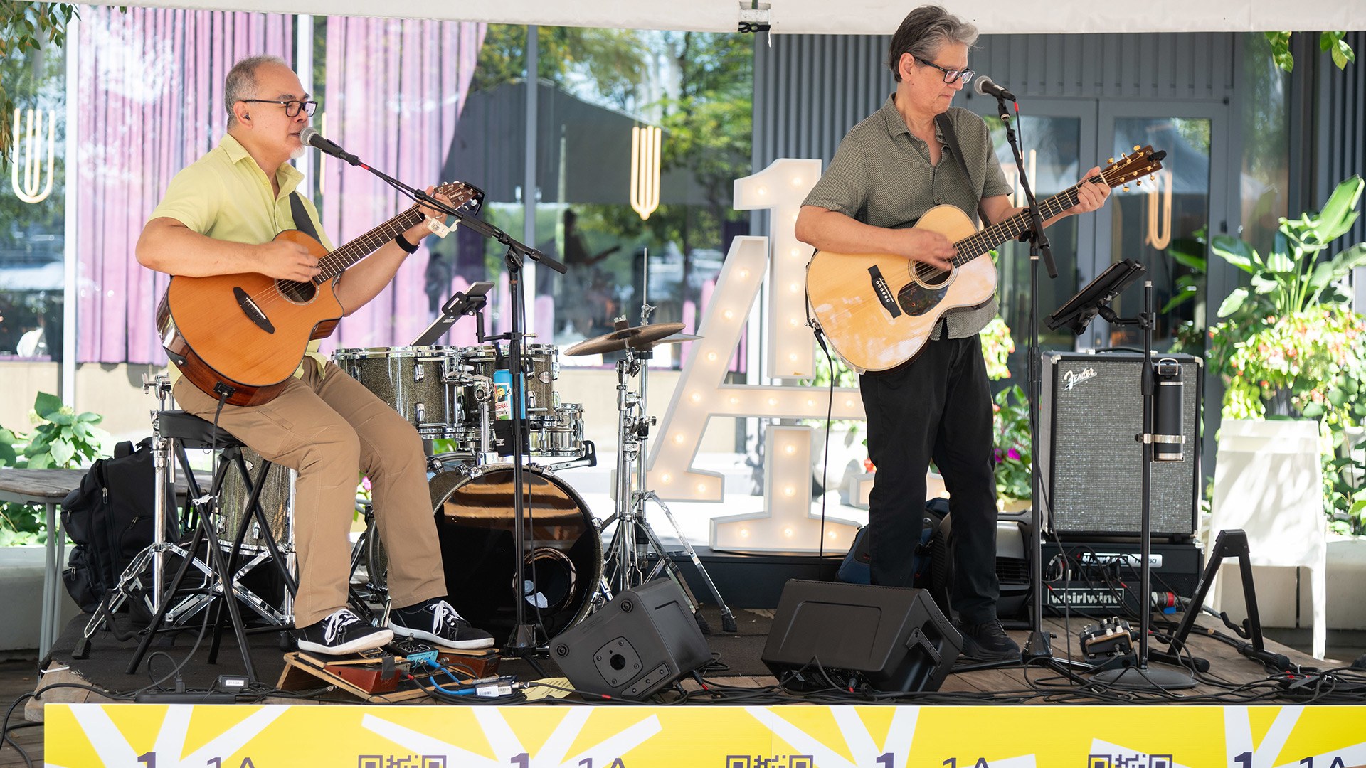 Two men, one seated and one standing, sing and play guitars on a stage in front of a lit-up 1A sign.