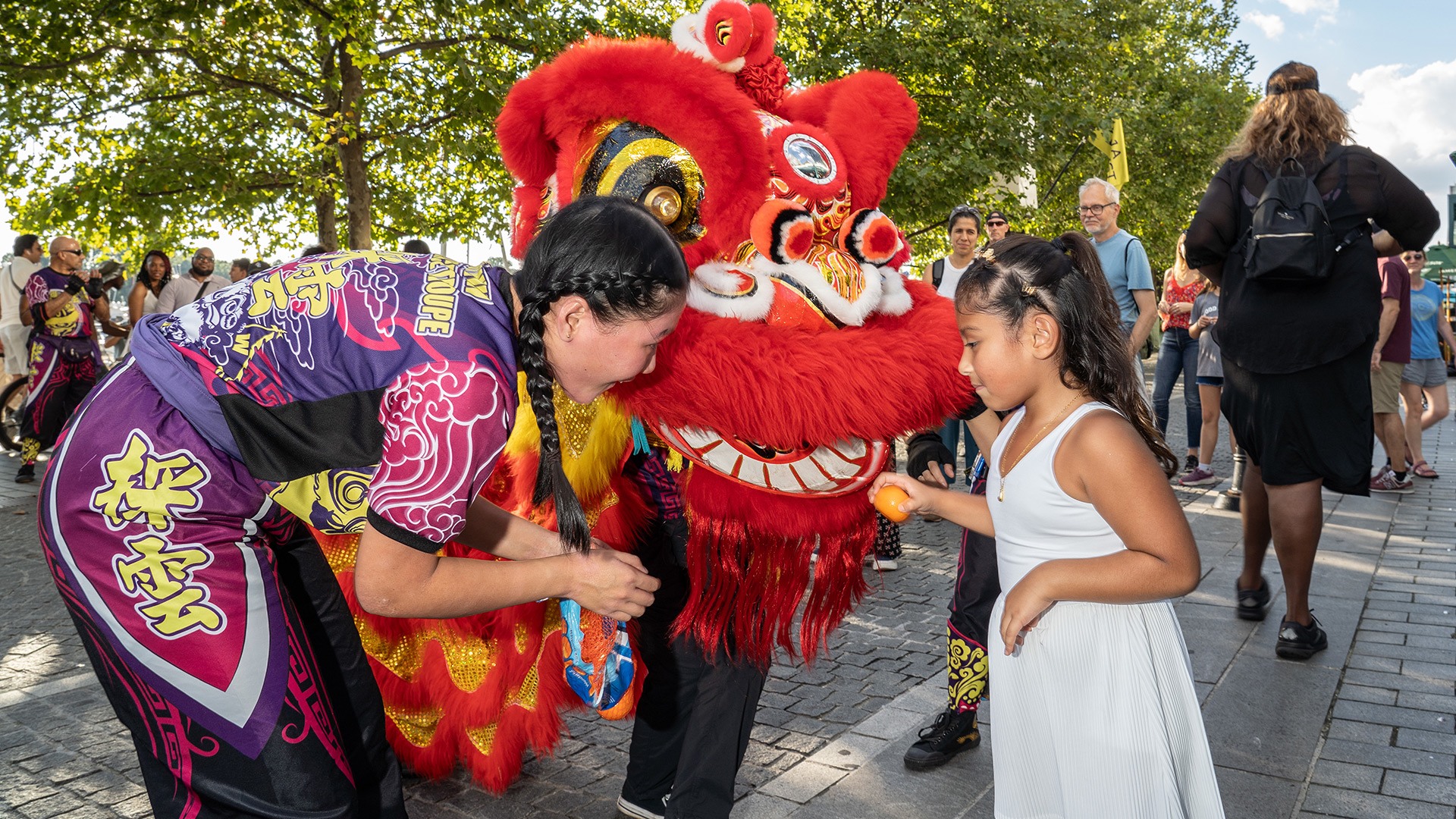 A woman with braided hair and a brightly colored costume covered in Chinese characters leans down to show a girl in a white dress a large, stylized lion head decorated with red fur.