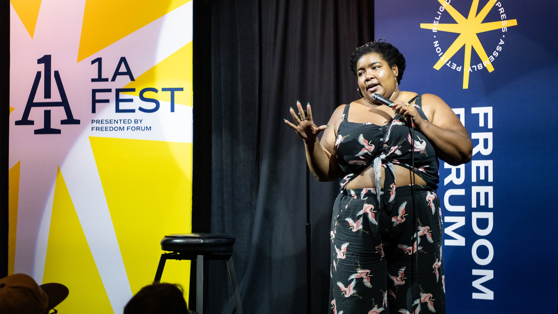 On a stage with 1A Fest signs, a woman with short, curly hair wearing a cropped, bird-print outfit stands with a microphone next to a stool and speaks to a crowd.