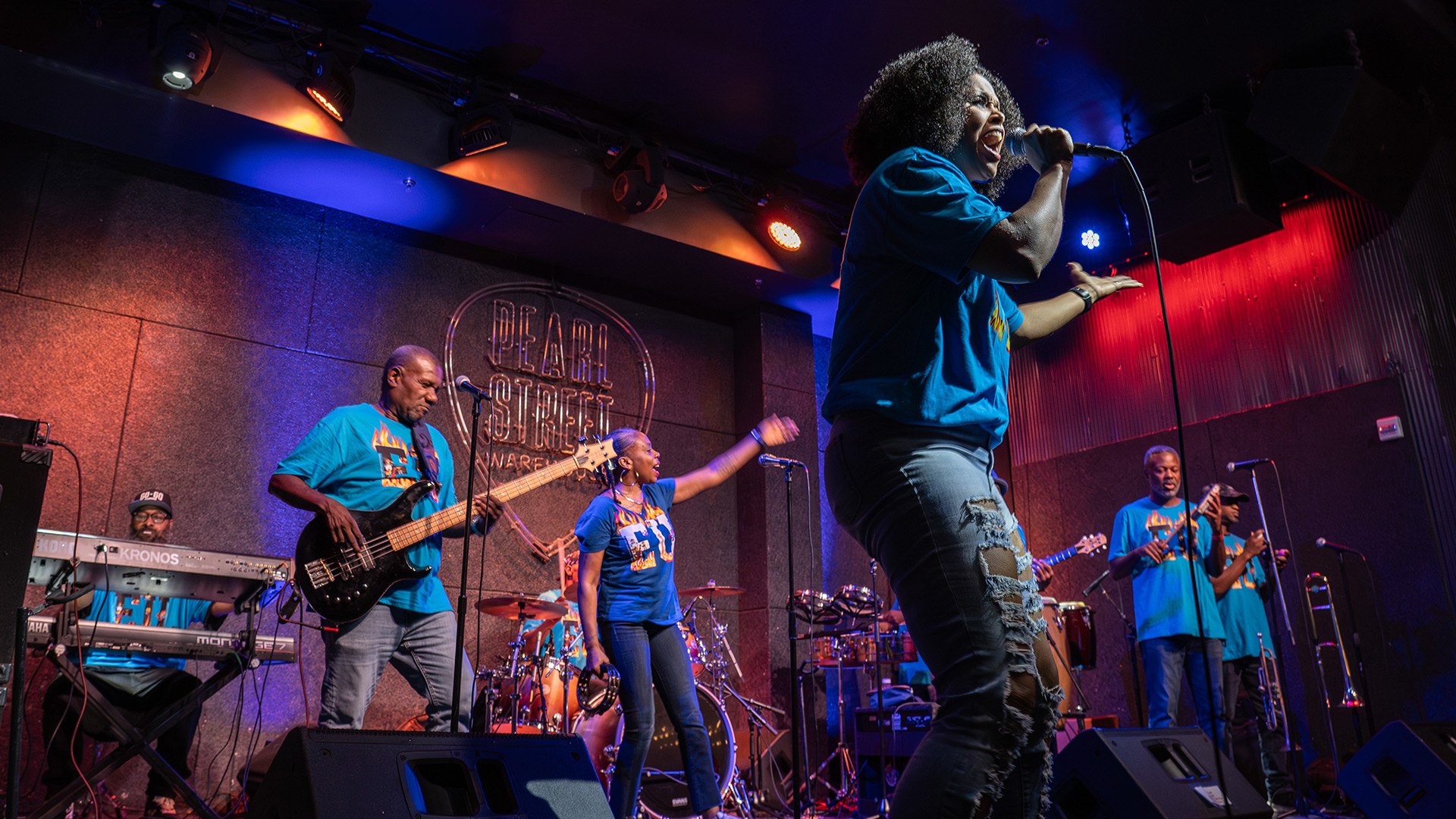 In front of a neon Pearl Street Warehouse sign, a band jams in shirts that say “EU” in letters ringed with flames. The band includes a keyboardist with a beard and ball cap, a bass guitarist, and two women singing enthusiastically.