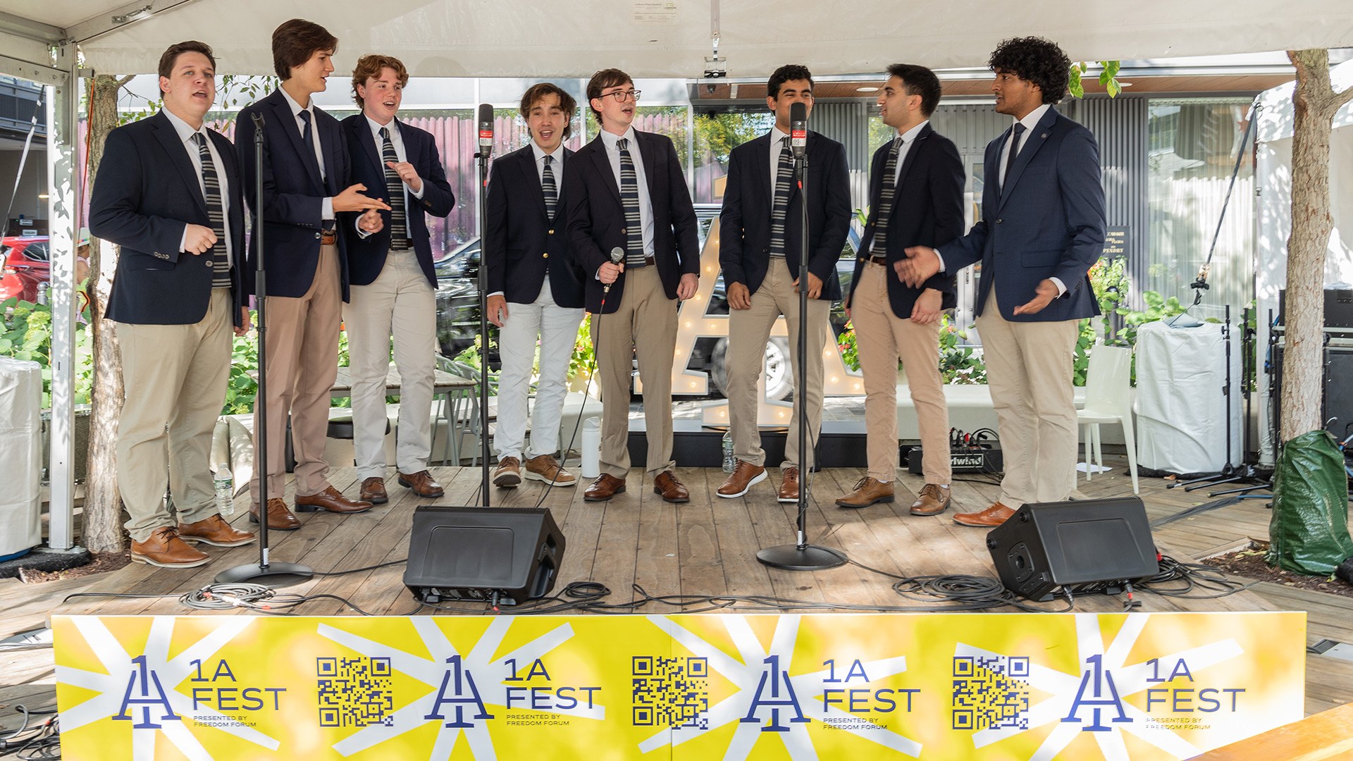 Eight young men in matching khaki slacks, dark jackets and striped ties sing acapella on a wooden stage.