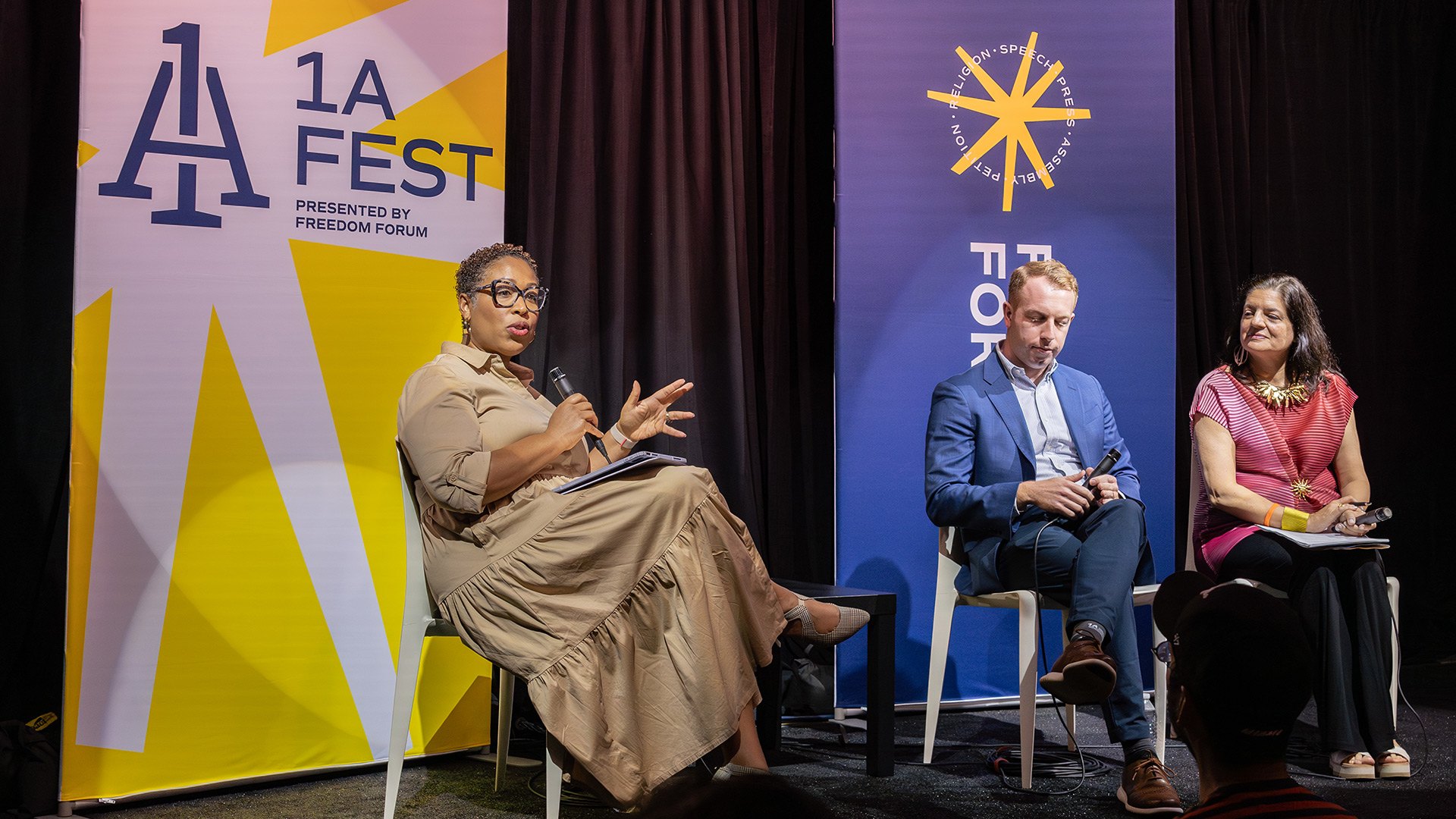 On a stage with 1A Fest signs, a woman with short, curly hair and glasses sits holding a microphone and talks to a man in a suit jacket and a woman with a gold bracelet also seated on stage.