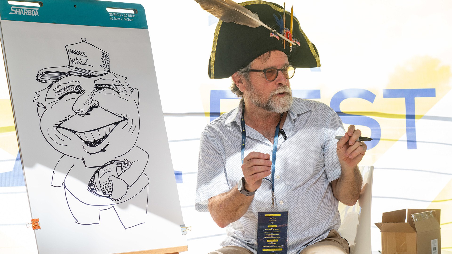 A man wearing a Napoleonic bicorne-style hat with gold trim and a feather sits next to an easel showing a caricature drawing.