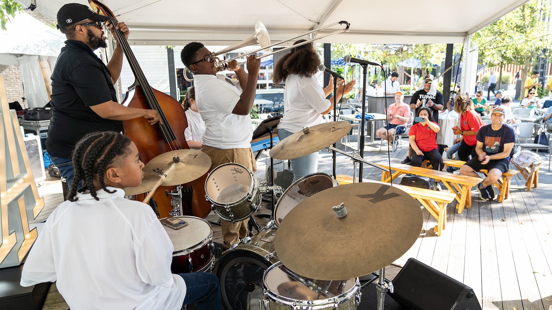 Guests sitting on benches look on as a jazz ensemble jams, including a young boy with braids on drums, a bearded man on upright bass, a boy with glasses on trombone, and a singing girl with curly pigtails.