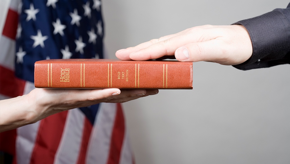 Close up of hand swearing on bible with american flag in background